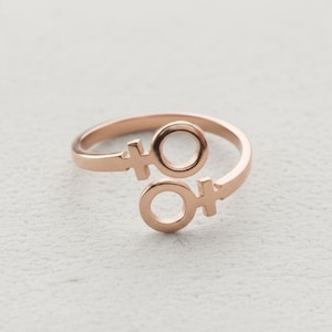 May include: Rose gold ring with two female gender symbols. The open design ring has a smooth, polished finish. The symbols are connected by a curved band, creating a modern and minimalist aesthetic. The ring is set against a neutral background.