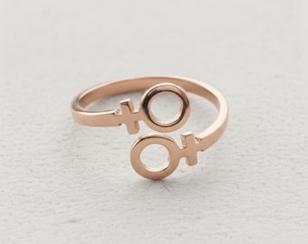 Puede incluir: Anillo de oro rosa con dos símbolos femeninos. El anillo de diseño abierto tiene un acabado liso y pulido. Los símbolos están conectados por una banda curva, creando una estética moderna y minimalista. El anillo está sobre un fondo neutro.