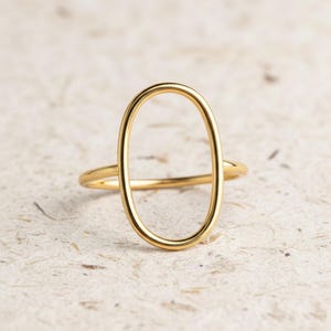May include: A gold ring with an open oval design. The ring has a slender band and a central, vertical oval. The background is a textured, neutral colour.