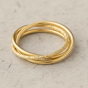 May include: A gold-colored ring with three interlocking bands. One band has a textured, dotted pattern, while the other two are smooth. The jewelry is displayed on a light gray surface.