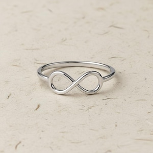 Peut inclure: Une bague argentée ornée d'un symbole infini. La bague est faite d'un métal brillant et réfléchissant, posée sur une surface texturée de couleur claire. Le symbole infini est l'élément central de la bague.