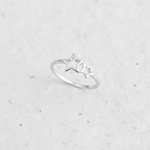Puede incluir: Un anillo plateado con dos estrellas caladas. El anillo tiene una banda delicada y un diseño minimalista. Las estrellas son de diferentes tamaños, una más grande que la otra, creando un tema celestial. El anillo probablemente está hecho de plata de ley.