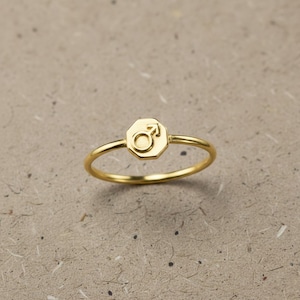 Könnte beinhalten: Ein goldfarbener Ring mit einem sechseckigen Emblem, das das Mars-Symbol trägt. Der Ring hat ein schmales Band und eine polierte Oberfläche. Dies ist ein Schmuckstück.