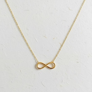May include: A gold-colored necklace featuring an infinity symbol pendant. The pendant is a continuous loop, suspended from a delicate chain. The necklace is displayed against a textured, off-white background.