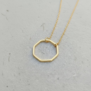 May include: A gold necklace with an octagonal pendant. The pendant is a geometric shape, suspended from a fine chain. The necklace is presented on a light grey surface, creating a minimalist aesthetic. The jewellery is simple and elegant.