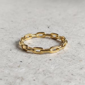 May include: A gold ring with a chain link design. The ring is made of a shiny, metallic material and features a series of interconnected links forming a continuous band. The ring is shown on a neutral background.