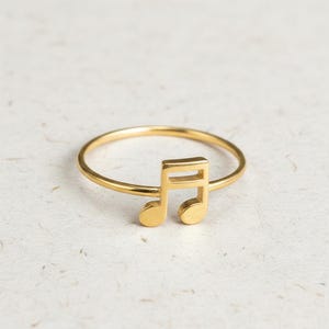 May include: A gold ring with a musical eighth note design. The ring is set against a textured, off-white background. The musical note is a prominent, three-dimensional element of the ring.