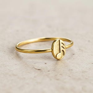 May include: A gold ring with a musical note motif. The ring has a slender band and a small, oval-shaped centrepiece displaying the musical note symbol. The ring is made of a shiny, metallic material.