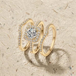 Peut inclure: Trois bagues en or, dont une bague de fiançailles avec un halo carré et une grosse pierre centrale, et deux alliances incurvées. Les bagues sont posées sur une surface texturée brun clair. Les bagues sont probablement en or et ornées de petites pierres scintillantes.