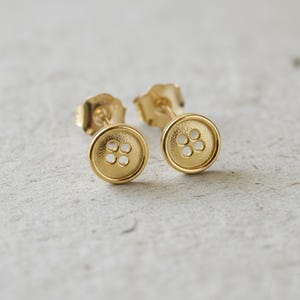 Puede incluir: Un par de pendientes de botón dorados. Cada pendiente presenta un diseño circular con cuatro pequeños agujeros en el centro, que se asemejan a un botón. Los pendientes tienen un poste y un respaldo para un uso seguro.