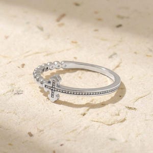 Puede incluir: Un anillo plateado con un diseño de cruz. El anillo tiene una textura de cuentas a lo largo de la banda y alrededor de la cruz. La cruz es el punto focal, con un diseño calado detallado. El anillo está sobre un fondo neutro.