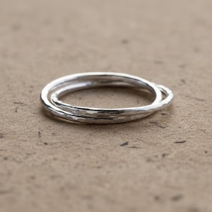Peut inclure: Ensemble de trois bagues argentées entrelacées avec une texture martelée. Les anneaux sont présentés sur une surface texturée brun clair. Bijoux simples et élégants.