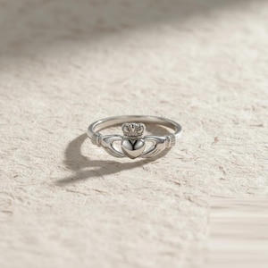 May include: A silver Claddagh ring with a heart, crown, and hands design. The heart is central, held by two hands, and crowned. The ring has a polished finish and is set against a textured, off-white background.