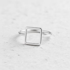 Könnte beinhalten: Ein minimalistischer Silberring mit einem hohlen quadratischen Design. Der Ring hat ein dünnes Band und eine polierte Oberfläche, wodurch eine moderne und geometrische Ästhetik entsteht. Der Ring wird auf einer strukturierten, hellfarbigen Oberfläche gezeigt.