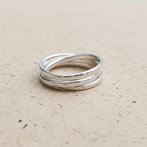 May include: A silver ring with a hammered texture, composed of three interwoven bands. The ring is presented on a neutral background, highlighting its simple and elegant design. This piece of jewellery is suitable for everyday wear.