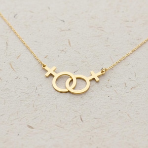 May include: A gold-colored necklace featuring two interlocking female symbols. The pendant is connected to a delicate chain. The necklace is displayed on a textured, off-white surface. The symbols are circular with a cross on top.