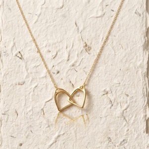 May include: A gold-colored necklace featuring two interlocking heart-shaped pendants. The delicate chain and pendants are made of a shiny, reflective metal. The necklace is displayed on a textured, off-white surface.
