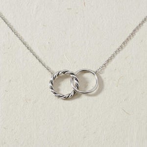 May include: A silver necklace with two intertwined circles. One circle has a twisted rope pattern, the other is a plain band. The necklace is suspended from a fine chain, offering a minimalist and elegant aesthetic.