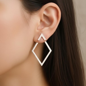 May include: Silver geometric earrings. The earrings feature a small triangle stud and a large open diamond shape. The earrings are worn on the ear, with the triangle stud positioned above the diamond.