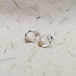 May include: A pair of silver stud earrings with a hammered finish. Each earring features a circular design with a small coil of gold-colored wire wrapped around the bottom. The earrings are displayed on a textured, off-white surface.