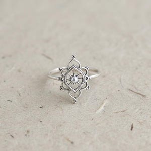 May include: A silver ring featuring an ornate, openwork design. The ring's focal point is a central starburst pattern, surrounded by a detailed, symmetrical floral-like motif. The ring is set against a neutral background, highlighting its intricate details.