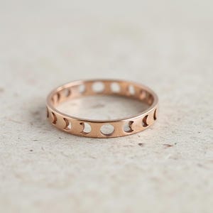 Puede incluir: Anillo de oro rosa con un diseño repetitivo de fases lunares. El anillo tiene una banda con formas de media luna recortadas y aberturas circulares, creando una estética delicada y celestial. El anillo es un estilo de banda simple.