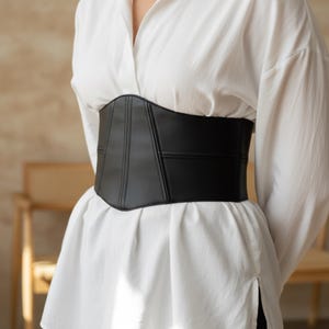 Può includere: Una cintura a corsetto nera dal design strutturato. La cintura è realizzata in un materiale liscio e presenta dettagli di cucitura verticali. È indossata sopra una camicia bianca abbottonata.