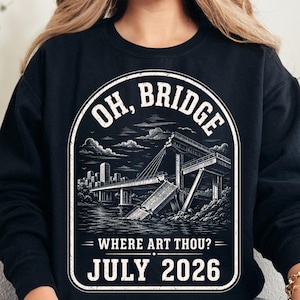 May include: A black long-sleeve sweatshirt featuring a white graphic print of a collapsed bridge over water with a city skyline in the background. The text reads "OH, BRIDGE WHERE ART THOU?" at the top and bottom, with "JULY 2026" printed at the base.