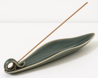 May include: A teal ceramic incense holder in a leaf shape with a curled handle. A brown incense stick is placed in the holder. The holder has a light beige rim and is set against a white background.