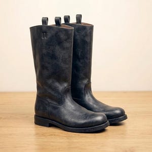 May include: A pair of black leather mid-calf boots with pull tabs at the top. The text FENDEX HANDMADE and a shield logo with the letter F are visible above the boots. The boots feature a rounded toe, a low heel, and visible stitching.