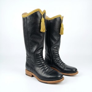 May include: A pair of black leather knee-high boots featuring a textured finish, a tan wooden heel and sole, and a decorative mustard yellow tassel attached to the top edge of each boot shaft, which is trimmed with matching yellow piping.
