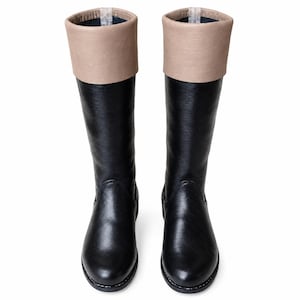 May include: A pair of knee-high boots featuring a smooth black lower section and a contrasting beige cuff at the top. The boots have a rounded toe, a flat sole, and a vertical pull tab visible at the inner top edge of each boot opening.