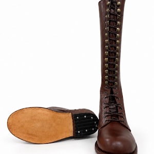 May include: A tall brown leather lace-up boot with brass eyelets and a dark brown lace. The boot features a rounded toe and a stacked heel. Beside it lies a second boot showing the tan leather sole with a black rubber heel insert.