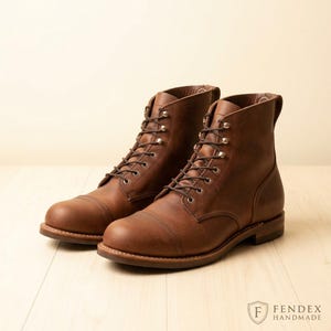 Puede incluir: Un par de botas de cuero marrón con cordones, diseño de puntera reforzada, ojales metálicos y cordones marrón oscuro. Las botas tienen un tacón apilado y una lengüeta en la parte trasera. El texto FENDEX HANDMADE es visible abajo a la derecha.