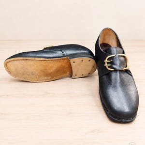 May include: A pair of black leather monk strap shoes featuring a gold-toned metal buckle closure. One shoe is positioned upright, while the other is placed on its side to reveal the tan leather sole and a stacked heel with visible metal nails.