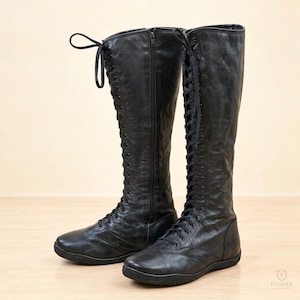 May include: A pair of black knee-high boots featuring a full-length lace-up front with black laces, a side zip closure, and a flat sole. The boots have a textured finish and a rounded toe design, extending to the calf with a structured shaft.