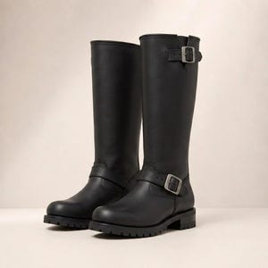 May include: A pair of black leather knee-high boots featuring a rounded toe, a thick lugged rubber sole, and a low block heel. Each boot includes an adjustable strap with a silver-toned metal buckle at the ankle and a second buckle at the top shaft.