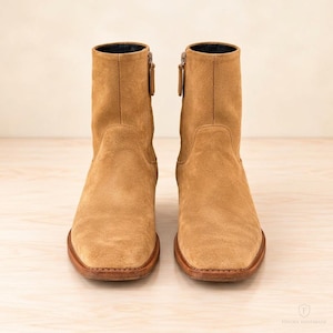 Könnte beinhalten: Ein Paar knöchelhohe Stiefel aus beigem Wildleder mit quadratischer Zehenpartie, seitlichem Reißverschluss mit passendem Zipper und einer braunen Ledersohle. Die Stiefel haben eine klare, strukturierte Form mit sichtbaren Nähten am Absatz.