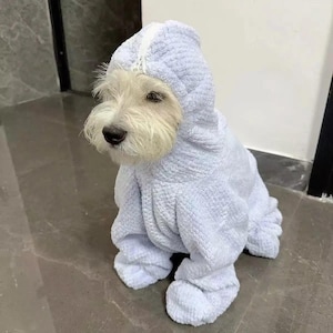 May include: A small, white dog wearing a light blue, hooded terrycloth outfit. The outfit covers the dog's body and legs, with a hood that covers the head. The dog is sitting on a grey tiled floor.