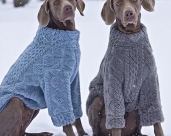 May include: Two Weimaraner dogs wearing matching cable-knit sweaters, one in light blue and the other in gray, sit in the snow. The dogs have brown fur and floppy ears. Snowflakes fall around them, creating a cozy winter scene.