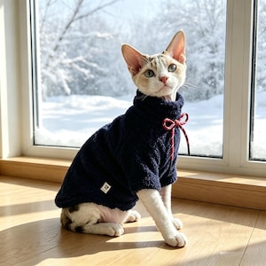Puede incluir: Un gato Sphynx lleva un suéter de forro polar azul marino oscuro con cuello alto y un cordón rojo y blanco en el cuello. El suéter presenta una pequeña etiqueta de marca rectangular blanca en el lateral inferior, con un tejido texturizado.