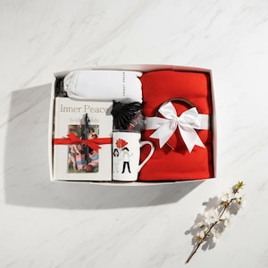 May include: A gift box with a red blanket tied with a white bow, a white mug with a couple graphic, a book titled "Inner Peace", a pen, a black hair accessory, and a white pillow.