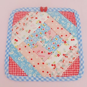 Peut inclure: Un dessous de plat carré et matelassé avec un motif patchwork. Le centre présente un mélange de tissus à motifs floraux, à pois et à cerises en rose, bleu et blanc. Une bordure en vichy bleu et des coins rouges en vichy encadrent le motif. Un petit nœud rouge est attaché en haut.