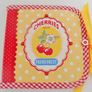 May include: A fabric-covered book with a yellow polka dot background and a red and white gingham border. The cover features a vintage-style label with the word "CHERRIES" and an illustration of cherries. A yellow ribbon with white polka dots is tied to the side.