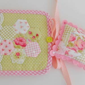 May include: A fabric notebook and pincushion set. The notebook has a green hexagon pattern with floral accents, a pink gingham border, and a lace edge. The pincushion is square with a pink scalloped edge and ribbon.
