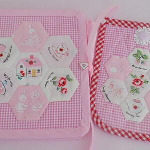 May include: A pink gingham fabric book and a matching rectangular pouch, both featuring white hexagonal patches with embroidered illustrations of houses, rabbits, flowers, and tea sets, accented with white lace trim and a small heart button.