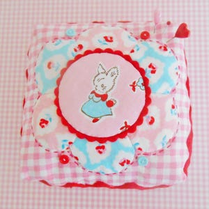 May include: A handmade pincushion with a floral design. The pincushion features a central pink circle with a cartoon bunny, surrounded by flower-shaped patches in pink and blue floral patterns. Red trim and buttons add detail.