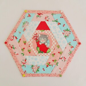 May include: A hexagonal patchwork quilt block with a pink, red, white, and blue color scheme. The center of the block features a vintage illustration of a cat wearing a red stocking and holding a candy cane. The block is surrounded by a pink border with white polka dots.