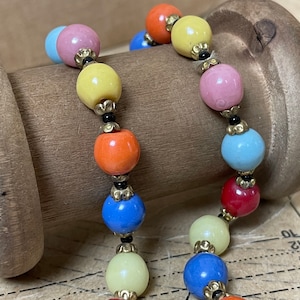 Könnte beinhalten: Eine bunte Perlenkette mit abwechselnd hellblauen, gelben, rosa, orangen und roten Perlen. Die Perlen sind durch kleine schwarze Perlen und goldene Akzente getrennt.