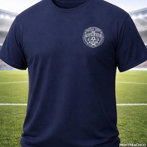 Puede incluir: Camiseta azul marino con un escudo con el texto "Tartan Army Scotland" y un gráfico de balón de fútbol. La camiseta es de manga corta y tiene cuello redondo. El fondo muestra un campo de fútbol y un estadio.